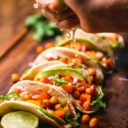 A person squeezes fresh ingredients onto colorful veggie tacos&mdash;chickpeas, peppers, herbs&mdash;while a lime sits nearby.