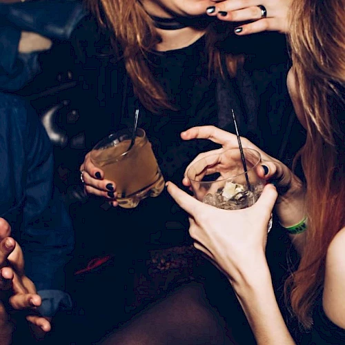 Two women in dark club lighting hold cups with straws, sipping smoky drinks while enjoying a night out.