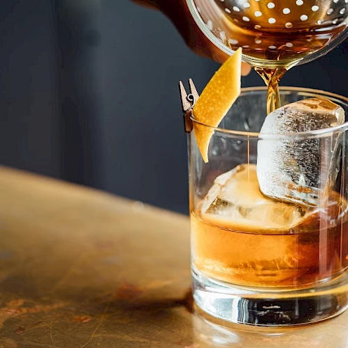 A bartender pours amber whiskey into an ice-filled rocks glass, garnished with an orange peel, for a classy cocktail.